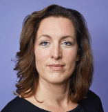 Eine Person mit schulterlangem braunem Haar blickt mit ruhigem Gesichtsausdruck direkt in die Kamera. Sie trägt ein schwarzes Oberteil und posiert vor einem violetten Hintergrund.