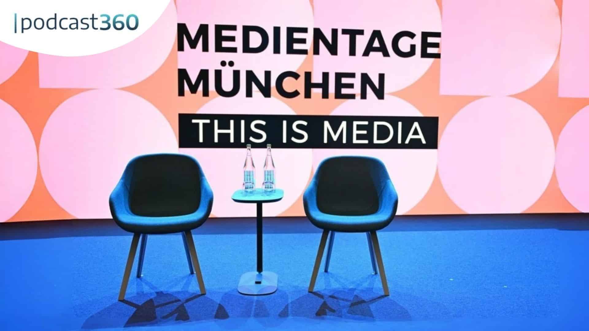 Zwei leere Stühle und ein kleiner Tisch auf einer Bühne mit "Medientage München: This Is Media" im Hintergrund.