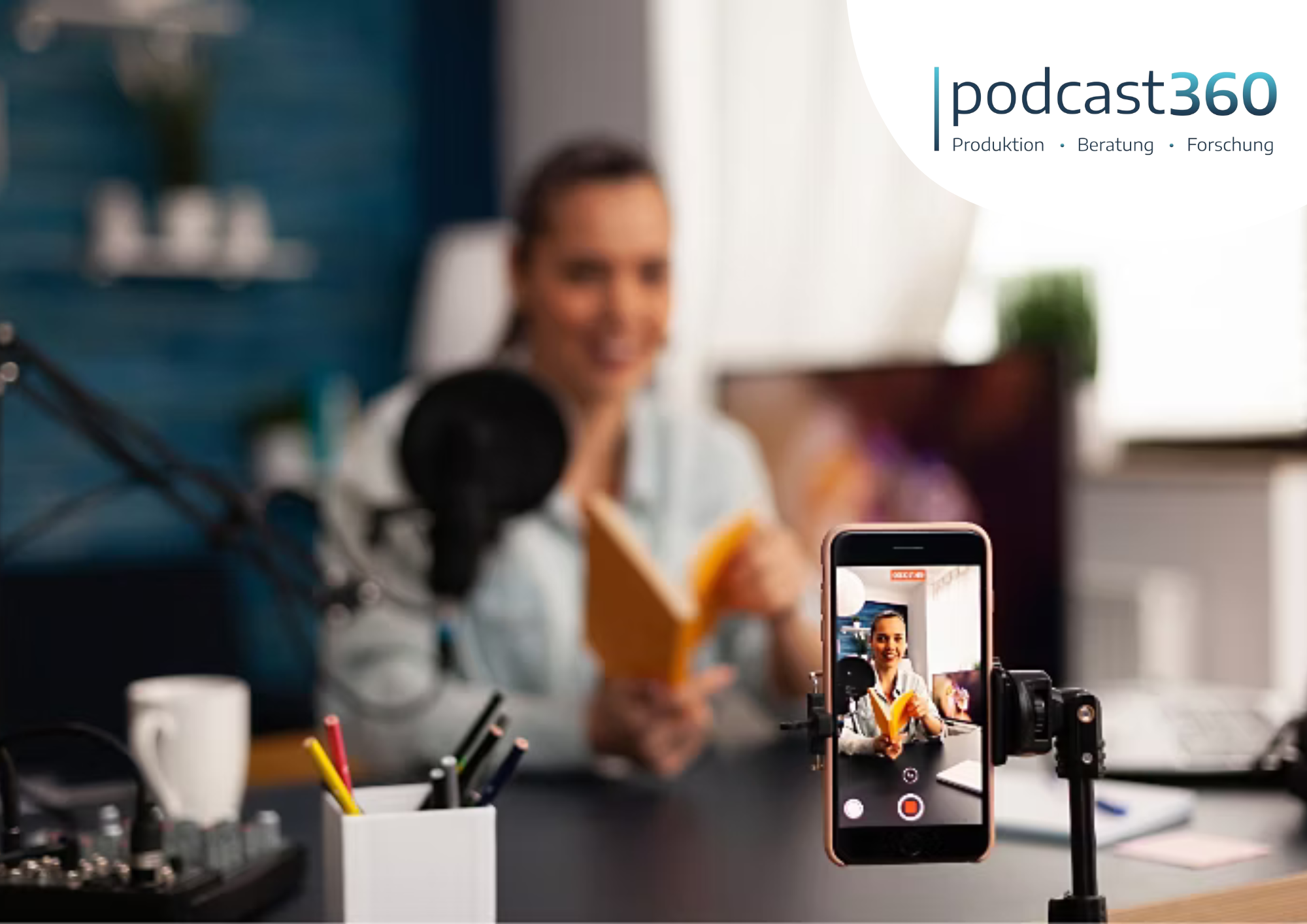 Eine Frau, die in einem Studio eine neue Podcasts-Folge aufnimmt, hält ein Buch in der Hand, während ihr Telefon filmt und das podcast360-Logo in der Ecke zu sehen ist.