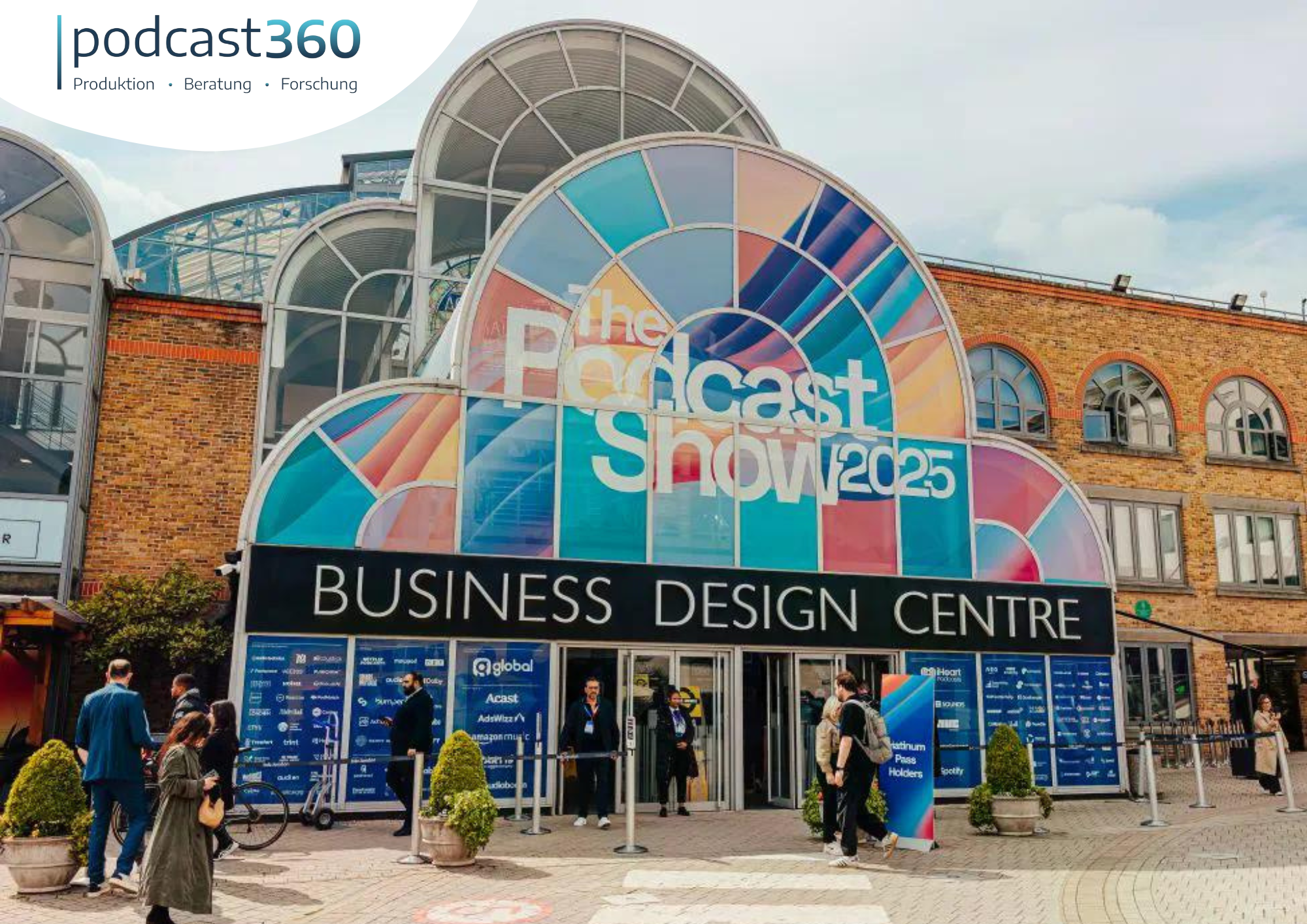 Eingang zur 2025 Podcast Show London im Business Design Centre, mit Menschen, die draußen spazieren gehen und lebhaftem Klickverhalten.