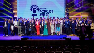 Eine große Gruppe posiert auf der Bühne mit Auszeichnungen beim Deutschen Podcast Award vor einem beleuchteten Schild.